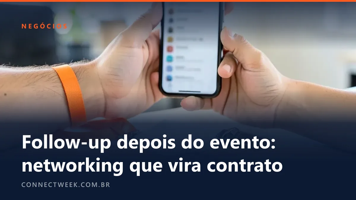 Mão segurando um celular com lista de contatos desfocada sobre uma pilha de cartões de visita em branco e um crachá na mesa